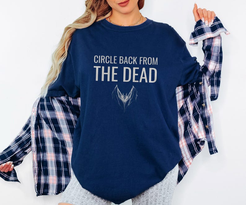 True Navy Comfort Colors t-shirt printed with the words Circle Back From The Dead and a drawing of a vintage silver paperclip bent into the shape of a bat, worn by a woman