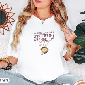 White Comfort Colors t-shirt printed with the words Mashed Potatoes Stuffing Cranberry Nap and a drawing of a heavy enamel-coated cast iron Dutch oven filled with creamy mashed potatoes, steam lines, worn by a woman