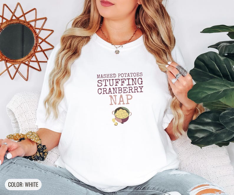 White Comfort Colors t-shirt printed with the words Mashed Potatoes Stuffing Cranberry Nap and a drawing of a heavy enamel-coated cast iron Dutch oven filled with creamy mashed potatoes, steam lines, worn by a woman