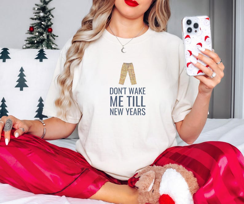 Ivory Comfort Colors t-shirt printed with the words Don'T Wake Me Till New Years and a drawing of a pair of plaid pajama pants folded neatly on a wooden stool, worn by a woman