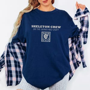 True Navy Comfort Colors t-shirt printed with the words Skeleton Crew On The Graveyard Shift and a drawing of a detailed medical defibrillator with its paddle cords forming a heart shape, flanked by t, worn by a woman
