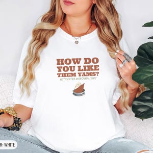 White Comfort Colors t-shirt printed with the words How Do You Like Them Yams? With Extra Marshmallows and a drawing of a roasted yam sliced in half with melted marshmallow topping, worn by a woman