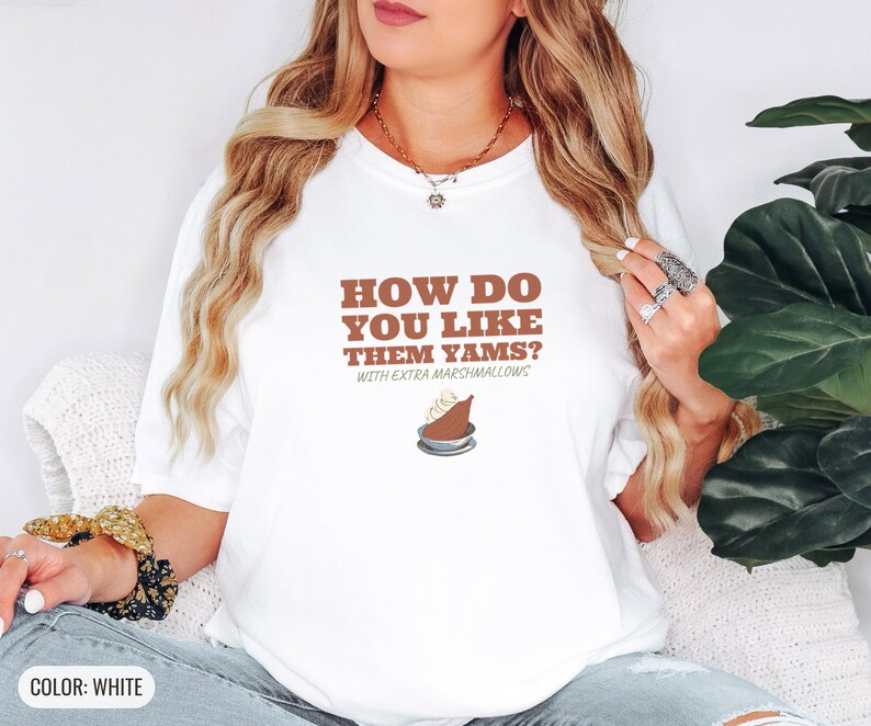 White Comfort Colors t-shirt printed with the words How Do You Like Them Yams? With Extra Marshmallows and a drawing of a roasted yam sliced in half with melted marshmallow topping, worn by a woman