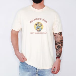Ivory Comfort Colors t-shirt printed with the words One Man'S Trash Is My Halloween Treat and a drawing of a scruffy opossum wearing a tiny plastic orange pumpkin trick-or-treat bucket on its head, worn by a man