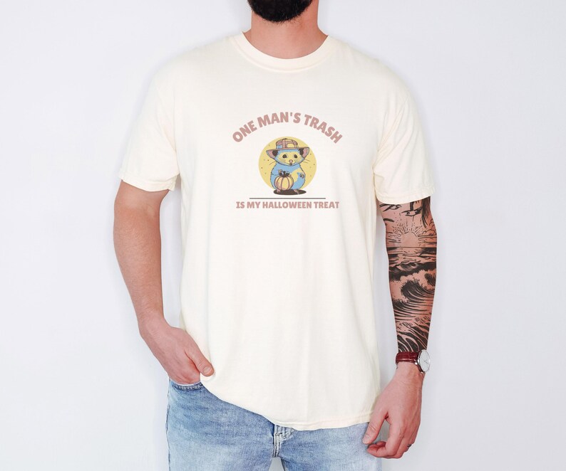 Ivory Comfort Colors t-shirt printed with the words One Man'S Trash Is My Halloween Treat and a drawing of a scruffy opossum wearing a tiny plastic orange pumpkin trick-or-treat bucket on its head, worn by a man