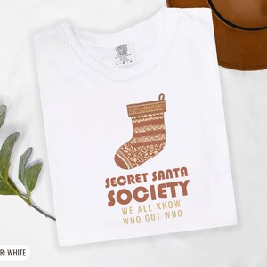 White Comfort Colors t-shirt printed with the words Secret Santa Society We All Know Who Got Who and a drawing of a knitted wool stocking stuffed with stick candy, folded flat lay