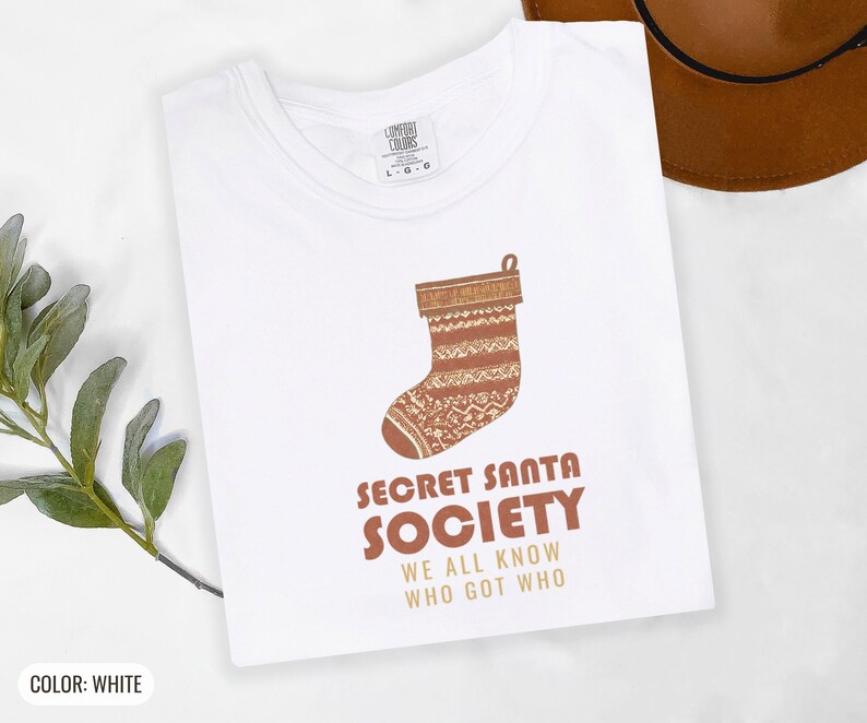 White Comfort Colors t-shirt printed with the words Secret Santa Society We All Know Who Got Who and a drawing of a knitted wool stocking stuffed with stick candy, folded flat lay