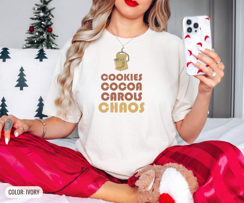 Ivory Comfort Colors t-shirt printed with the words Cookies Cocoa Carols Chaos and a drawing of a retro metal hand mixer with steel beaters, worn by a woman