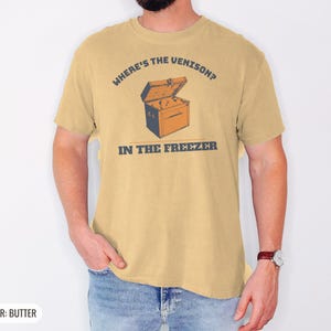 Butter Comfort Colors t-shirt printed with the words Where'S The Venison? In The Freezer and a drawing of a vintage metal chest freezer with a pair of paper-wrapped butcher packages resting on top, worn by a man