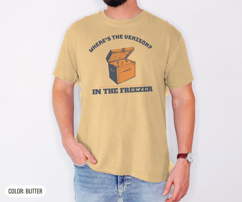 Butter Comfort Colors t-shirt printed with the words Where'S The Venison? In The Freezer and a drawing of a vintage metal chest freezer with a pair of paper-wrapped butcher packages resting on top, worn by a man