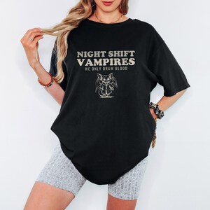 Black Comfort Colors t-shirt printed with the words Night Shift Vampires We Only Draw Blood and a drawing of a cute cartoon vampire bat wearing a tiny nurse scrub top, worn by a woman