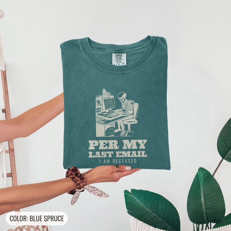 Blue Spruce Comfort Colors t-shirt printed with the words Per My Last Email I Am Deceased and a drawing of a skeleton sitting at an office desk typing on a computer keyboard, folded flat lay