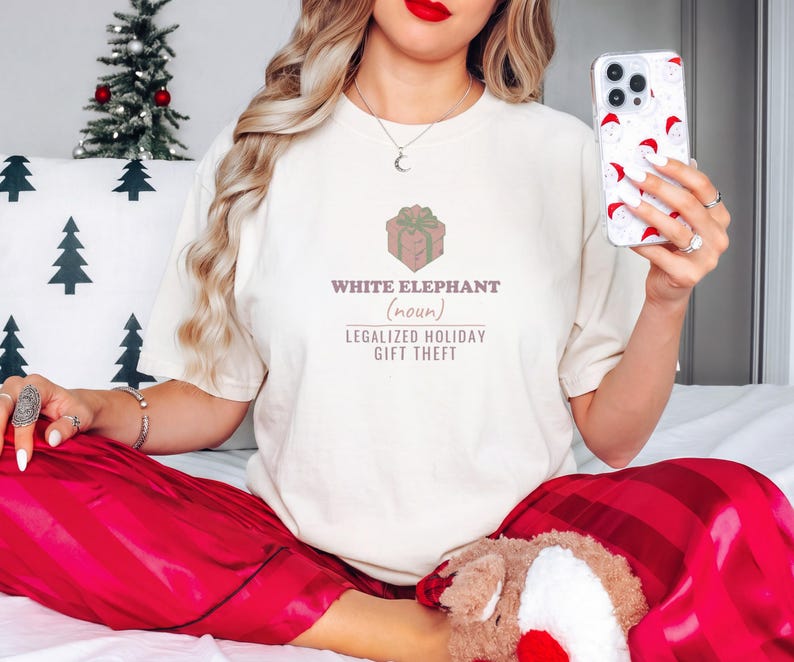 Ivory Comfort Colors t-shirt printed with the words White Elephant (Noun) Legalized Holiday Gift Theft and a drawing of a shiny wrapped gift parcel with a giant comical polka-dot bow, worn by a woman