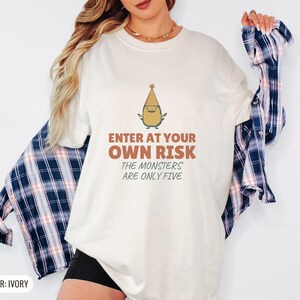 Ivory Comfort Colors t-shirt printed with the words Enter At Your Own Risk The Monsters Are Only Five and a drawing of a cute, smiling little green monster wearing a tiny paper party hat and holding a crayon, worn by a woman