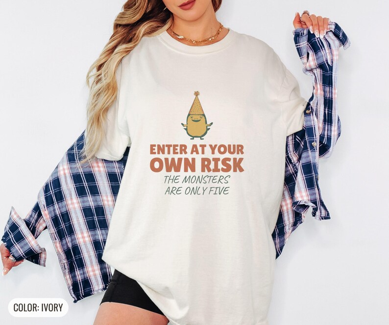 Ivory Comfort Colors t-shirt printed with the words Enter At Your Own Risk The Monsters Are Only Five and a drawing of a cute, smiling little green monster wearing a tiny paper party hat and holding a crayon, worn by a woman