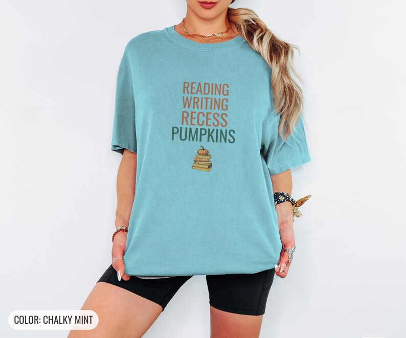 Chalky Mint Comfort Colors t-shirt printed with the words Reading Writing Recess Pumpkins and a drawing of a stack of three vintage schoolbooks with a small orange pumpkin sitting on top, worn by a woman