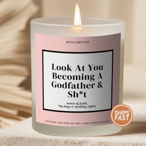 May include: A frosted glass candle with a lit wick and a pink label. The label reads: JOYLUMINATE, Look At You Becoming A Godfather & Sh*t, notes of faith, big hugs & spoiling rights, 8 OZ VEGAN, 100% PURE SOY WAX, HAND POURED IN THE USA.