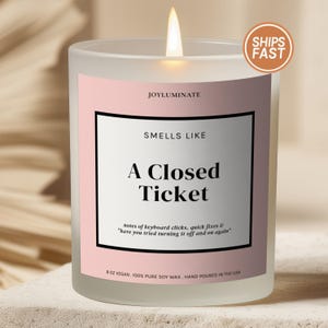 May include: A frosted glass candle with a pink label featuring the text JOYLUMINATE, SMELLS LIKE A Closed Ticket, and notes of keyboard clicks, quick fixes & have you tried turning it off and on again. The candle contains 8 oz of soy wax.