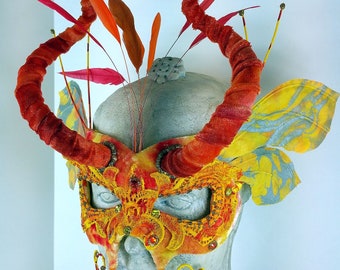 May include: A masquerade mask with orange, yellow, and red fabric and feathers. The mask has a whimsical design with intricate details and a dramatic, curved shape.