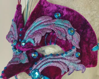 May include: A masquerade mask made of pink velvet with blue and silver embroidery and rhinestones. The mask has a long, pointed nose and is decorated with blue feathers.