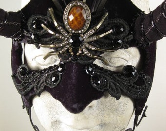 Puede incluir: Una máscara de terciopelo negro para mascarada con un diseño de araña y una gran joya de ámbar en el centro. La máscara tiene plumas negras en la parte superior y un ribete de encaje negro alrededor de los bordes.