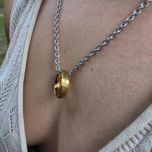 May include: A gold-colored metal ring featuring engraved script text around its outer surface, suspended from a silver-colored metal chain necklace. The ring hangs vertically from the chain, displaying a polished finish and detailed etched markings.