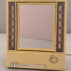 May include: A beige Clairol True-to-Light makeup mirror with a rectangular frame, a central pivoting mirror, and two vertical columns of five circular light bulbs. The base features a power switch, a dial for light settings, and a 120V 60Hz 1500W outlet.