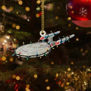 May include: A flat, starship-shaped Christmas tree ornament featuring the NCC-1701-E designation. The silver-toned vessel is wrapped in a string of miniature, multi-colored LED lights and hangs from a gold-colored cord attached to a circular loop.