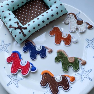 May include: A collection of six embroidered horse patches in red, blue, dark brown, light brown, green, and white, each featuring a saddle and tail. The patches are arranged on a white plate with blue starfish patterns near a mint green and brown star-print fabric cushion.