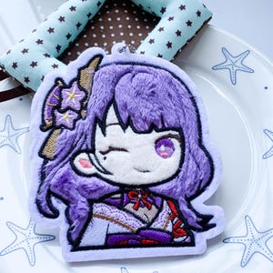 May include: A plush keyring featuring an embroidered anime-style character with vibrant purple hair, a winking expression, and a detailed kimono outfit. The character has a small flower accessory in their hair and is rendered in a soft, textured fabric.