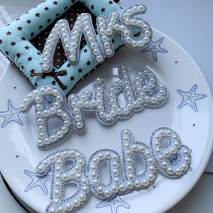 May include: Three white fabric patches featuring the words Mrs, Bride, and Babe in cursive script. Each letter is outlined with small clear rhinestones and filled with white faux pearls, creating a textured, dimensional appearance on the surface.