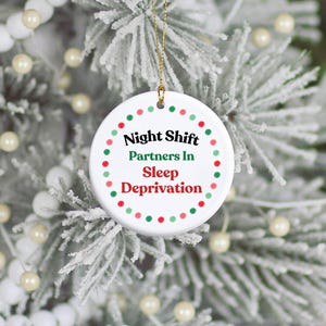 May include: A round white ceramic ornament featuring a gold hanging string. The text reads Night Shift Partners In Sleep Deprivation in black, green, and red lettering, surrounded by a circular border of alternating red and green dots.