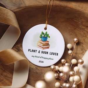 May include: A round white ceramic ornament hanging by a gold string. The center features a watercolor illustration of a potted green plant atop a stack of books. Text reads: PLANT & BOOK LOVER, my kind of happy place, and 2026.