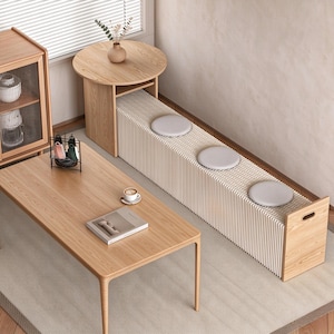 May include: A light wood coffee table, a matching round side table, and a long, expandable accordion-style bench with a white pleated structure and three round grey cushions. A wooden cabinet with glass doors and a book are also present.