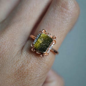 May include: A rose gold ring featuring a rectangular, textured olive-green stone at the center. The setting is adorned with intricate scrollwork and small, clear accent stones along the sides of the central gemstone.