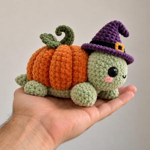 May include: A crocheted turtle figurine featuring a bright orange pumpkin shell with green leaves and a curly stem. The turtle has a light green body, black plastic eyes, pink cheeks, and wears a purple witch hat with a black band and orange buckle.