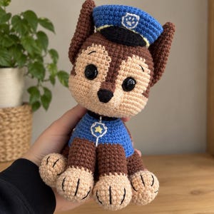 May include: A brown and tan crochet plush toy of a dog character wearing a blue police uniform vest with a yellow star emblem and a blue police cap featuring a white paw print badge. The toy has black plastic eyes and a black nose.