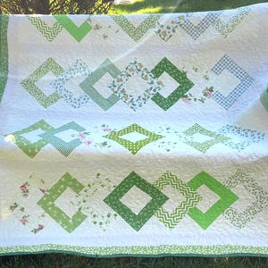 May include: A white quilt featuring a repeating pattern of green diamond-shaped frames. The diamonds are composed of various fabrics in shades of green, including gingham, floral, and chevron prints. The quilt has a green and white gingham border.