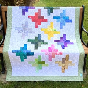May include: A quilt featuring a white background with a swirling stitched pattern and multiple cross-shaped blocks in various colors including red, blue, yellow, green, purple, pink, and tan, finished with a light green border.