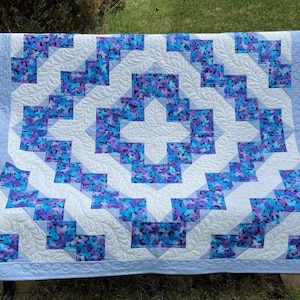 May include: A quilt featuring a geometric pattern of blue and purple butterfly-print fabric squares alternating with white fabric strips. The white sections are quilted with a feather design, and the border is a solid light blue fabric.