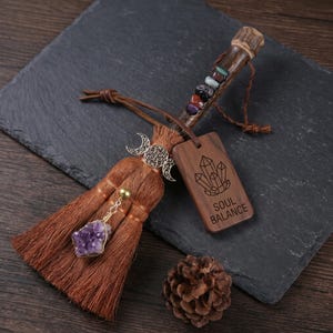 Puede incluir: Una escoba decorativa con cerdas marrones, un mango de madera con siete pequeñas piedras de colores, un dije de luna triple plateado y un cristal de amatista morado colgante. Una etiqueta de madera rectangular tiene grabado SOUL BALANCE y cristales.