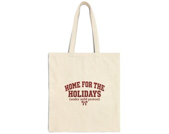 Puede incluir: Un bolso de tela color crema con dos asas para el hombro. La parte frontal presenta un texto en rojo oscuro que dice "HOME FOR THE HOLIDAYS (under mild protest)" centrado sobre un pequeño gráfico simple de un lazo.