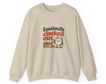 Puede incluir: Una sudadera beige de cuello redondo con un gráfico de un despertador cansado sosteniendo una taza de café. El texto "Emotionally clocked out since 9:02 AM" aparece en letras de color verde oscuro y rojo en el centro de la prenda.