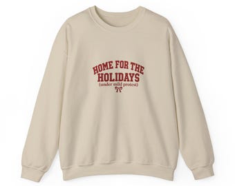 Puede incluir: Una sudadera de cuello redondo en color beige con el texto en rojo oscuro "HOME FOR THE HOLIDAYS (under mild protest)" impreso en el pecho, acompañado de un pequeño gráfico de un lazo rojo centrado bajo el texto.