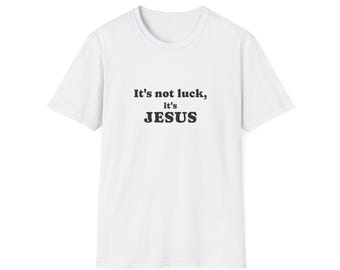 Puede incluir: Una camiseta blanca de manga corta con texto negro impreso en el centro del pecho. El texto dice, It's not luck, it's JESUS, en una fuente serif en negrita. La prenda tiene un cuello redondo clásico y un dobladillo recto estándar.