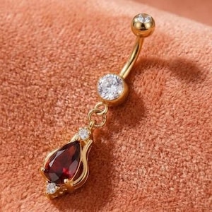 May include: A gold-toned navel ring featuring a curved barbell with a round clear stone at the top and a dangling teardrop-shaped deep red stone at the bottom, accented by two small clear stones flanking the teardrop setting.