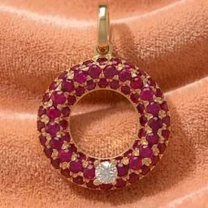 May include: A circular gold pendant featuring a pavé setting of numerous deep red rubies covering the entire ring surface, with a single clear, round-cut diamond set at the bottom center and a polished gold bail at the top for attachment.