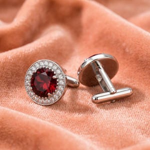 May include: A pair of silver-toned cufflinks featuring a round, deep red faceted center stone surrounded by a halo of small, clear, sparkling stones. The cufflinks have a polished metallic finish and a standard T-bar toggle closure mechanism.