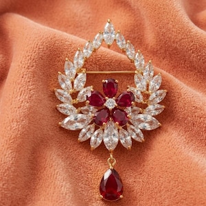 May include: A gold-toned brooch featuring a central flower design with five deep red, pear-shaped stones. The flower is surrounded by a wreath of clear, marquise-cut stones, with a single, dangling, pear-shaped red stone at the bottom.