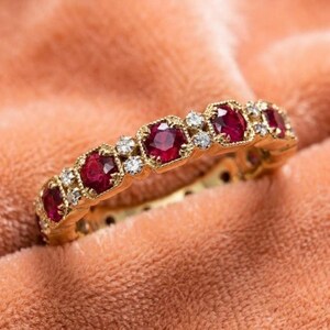 May include: A yellow gold band ring featuring a repeating pattern of round faceted red rubies and small clear white diamonds. The metal setting has a textured, beaded edge surrounding each gemstone, creating a continuous, detailed design.